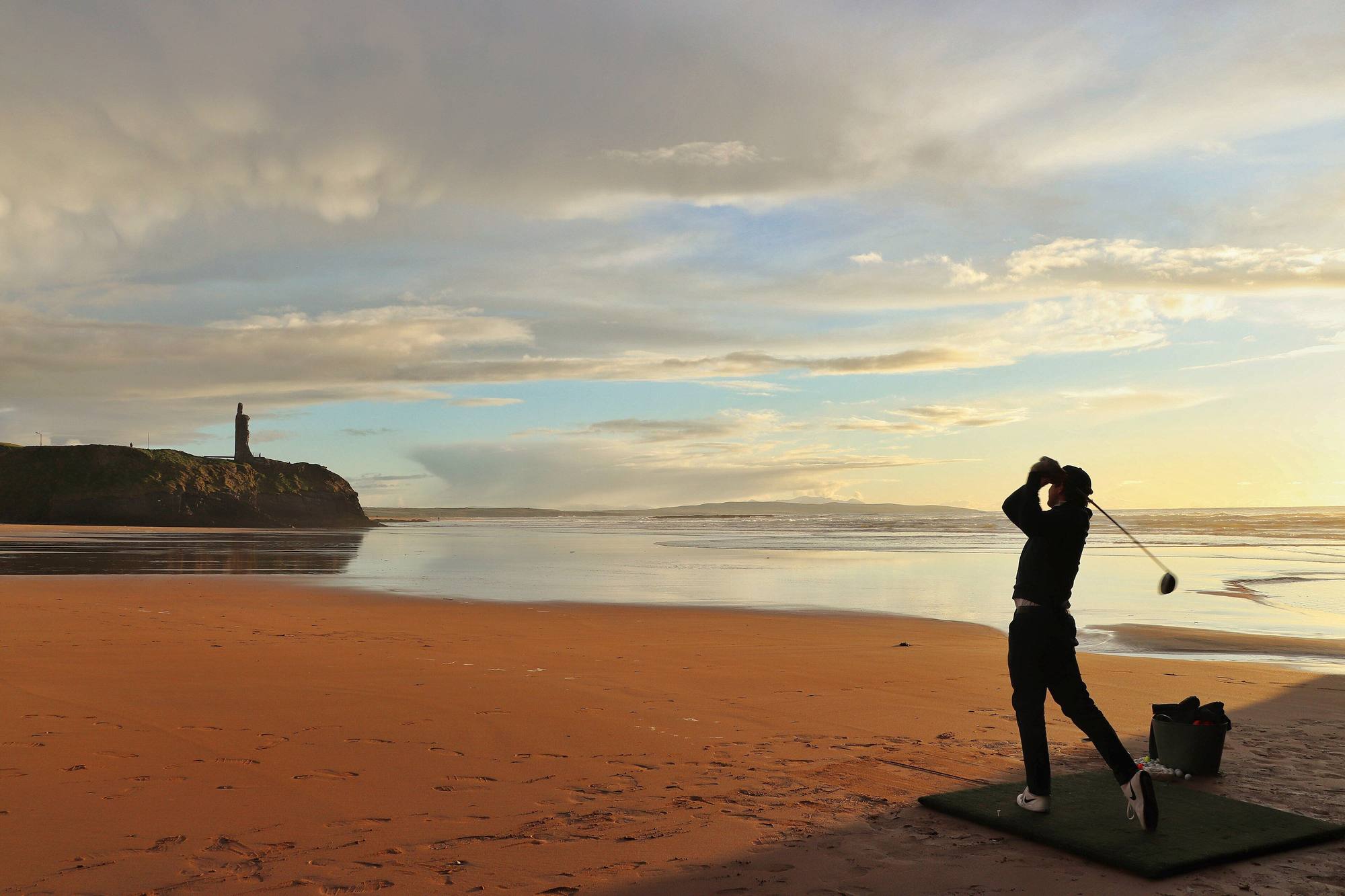 A golfer drives off a matt on a wide strand at sunset, a ruined tower on the headland behind